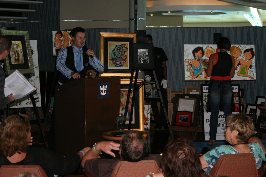 Wayne auctioneering at a Royal Caribbean art auction with Natalie Dyer live painting behind him and her work waiting to go under the hammer