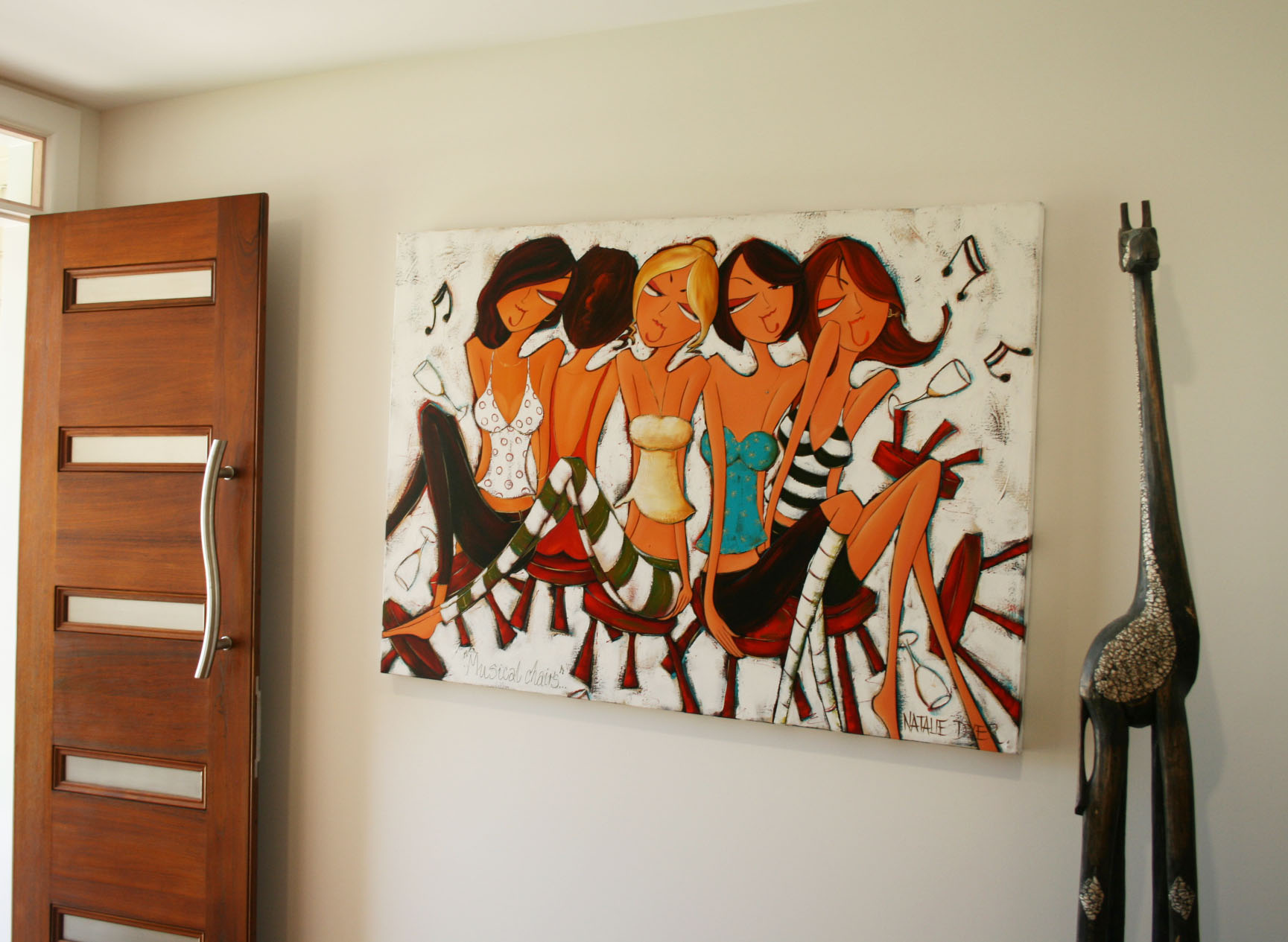 Musical Chairs hanging beside the timber front door of a collector's home, with a tall carved wooden giraffe standing alongside it